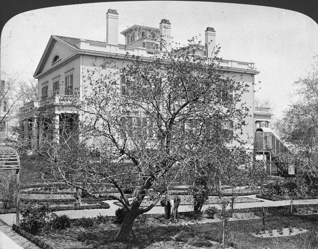 1870s photo of a branching apple tree and garden plots beside large, two story rectangular house