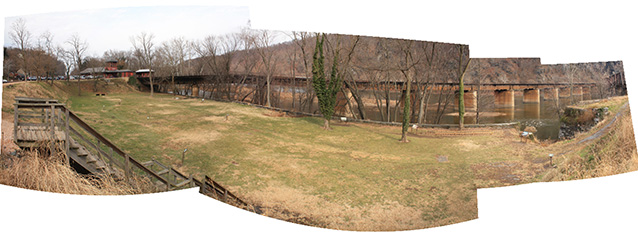 Stitched panorama shows a wide view of a level, grassy area between a berm and the river.