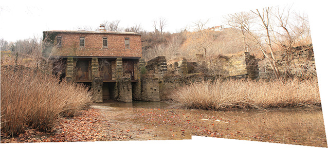 The stone and brick remains of a mill stand along a canal with shallow water and leafless shrubs.