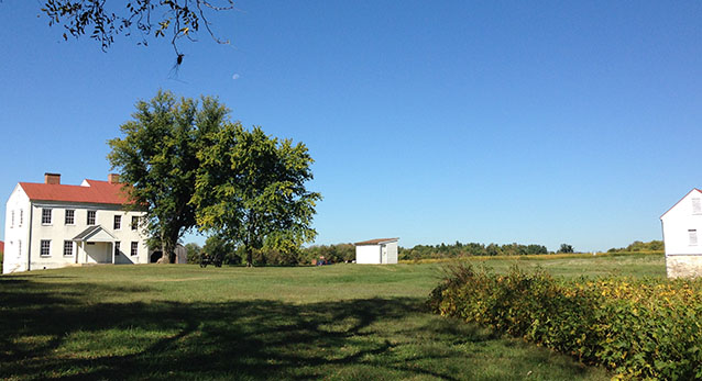 A two-story farm house beside a tree, in an open agricultural landscape with fields and outbuildings