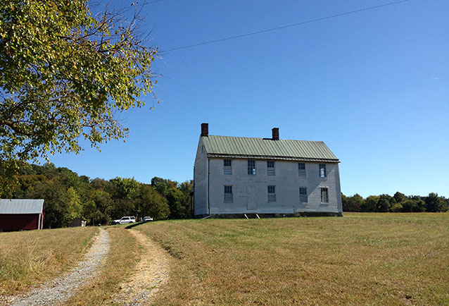 Dirt tire tracks curve toward a two-story farmhouse under clear sky.