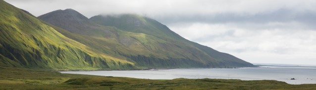A coastal bay leads to green slopes of a mountain range topped by thick fog.