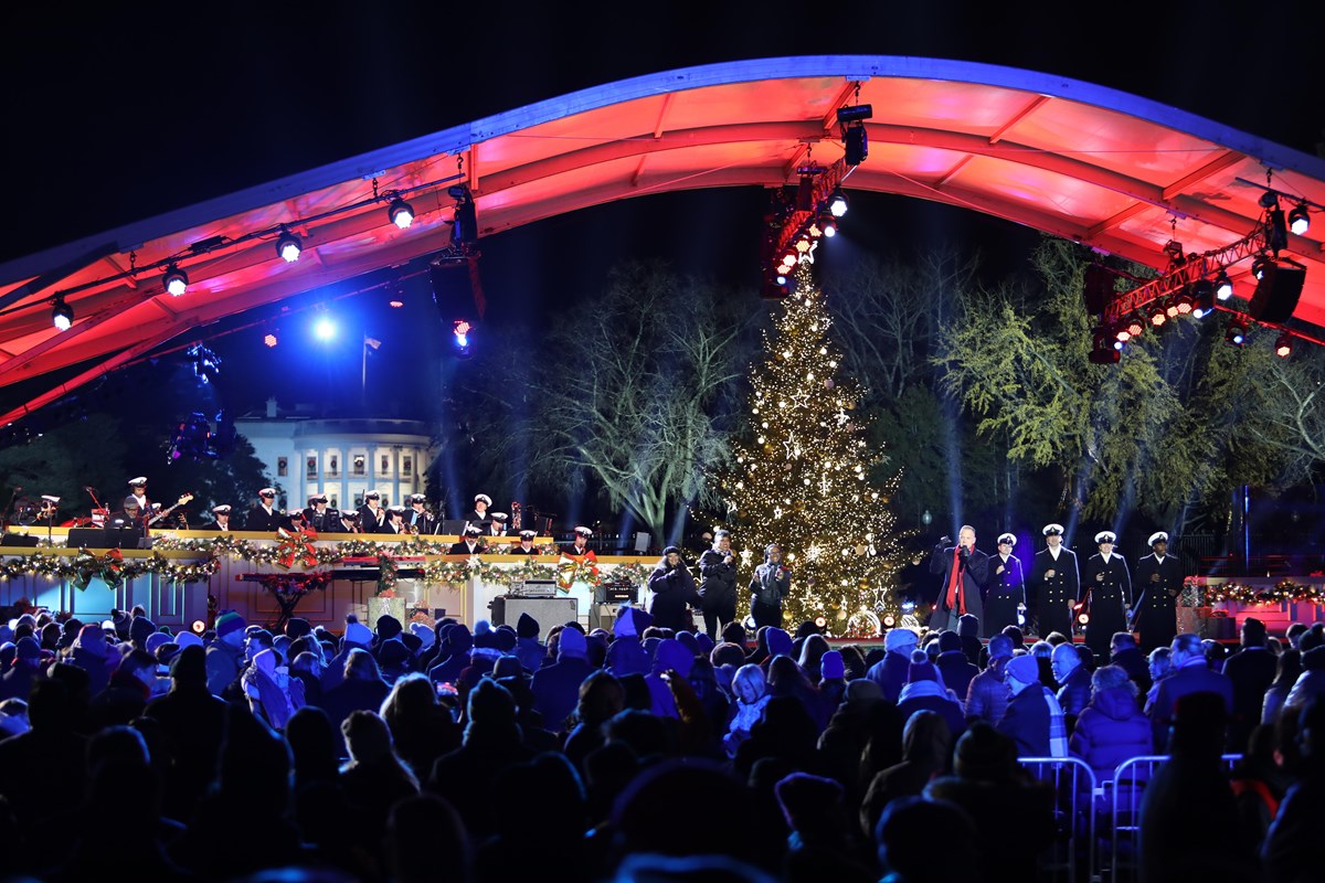 Crowds in foreground watch a performance on the stage before the National Christmas Tree at night.