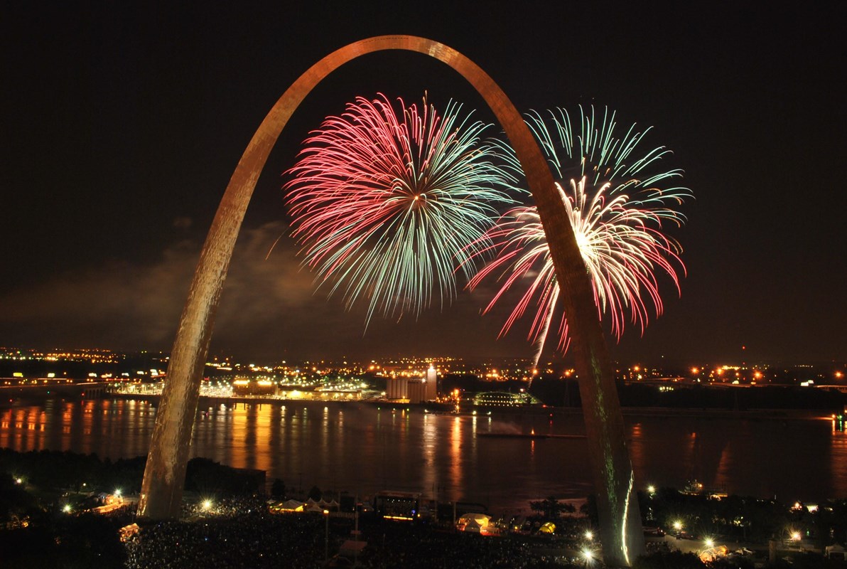 red, white and blue fireworks behind the Gateway Arch with lights reflecting in the river