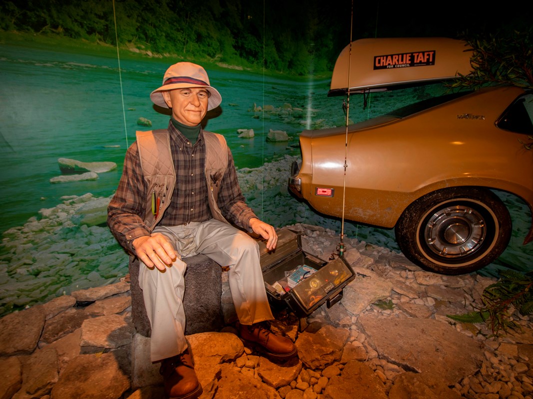 A sitting model of an older man in a vest, shirt and pants next to a replica of a car & canoe on top