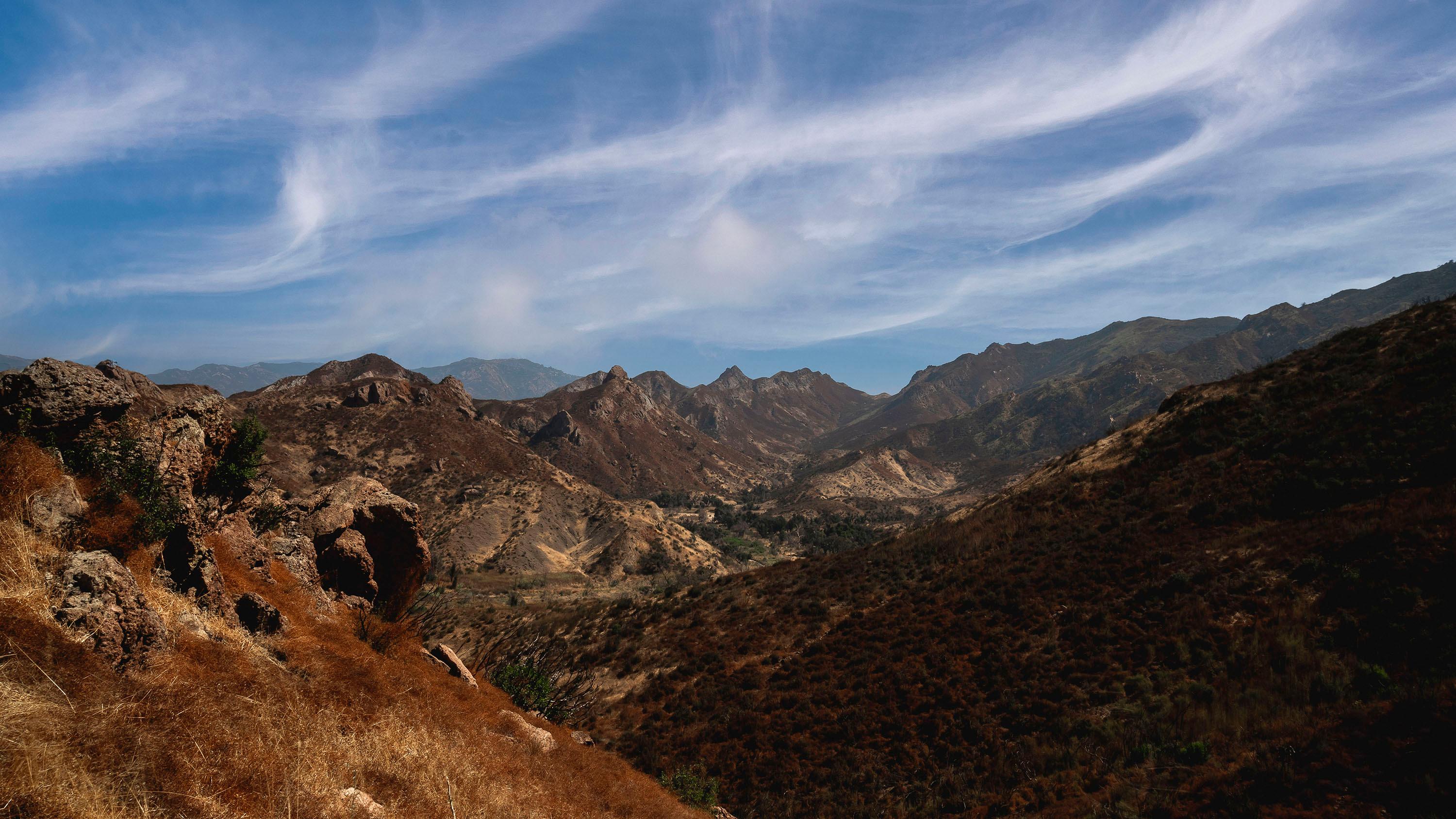 Wispy cloud covered sky over chaparral covered mountains.