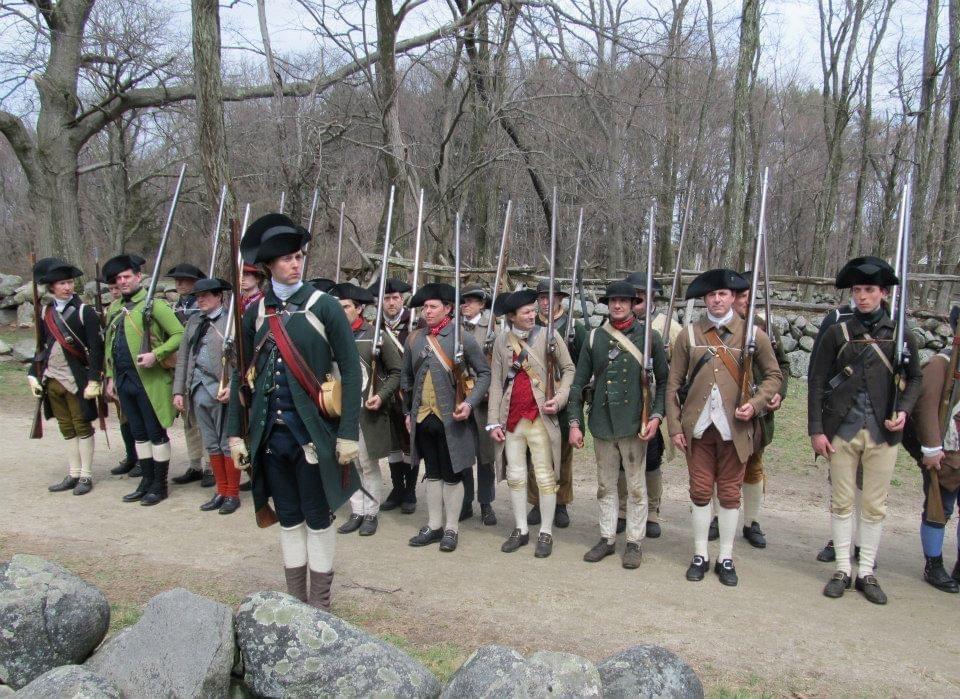 A company of armed minute men stand in formation along a dirt road with the officer out in front.