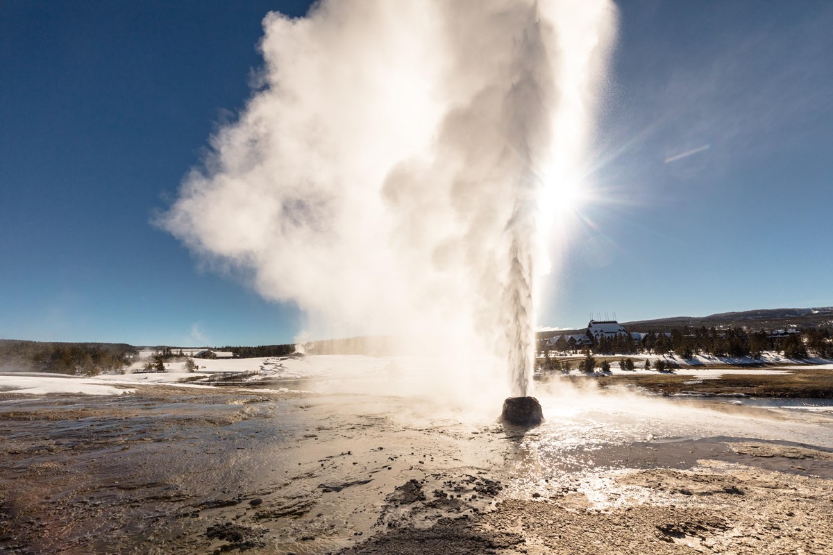 A geyser erupts steam in front of the bright sun. 