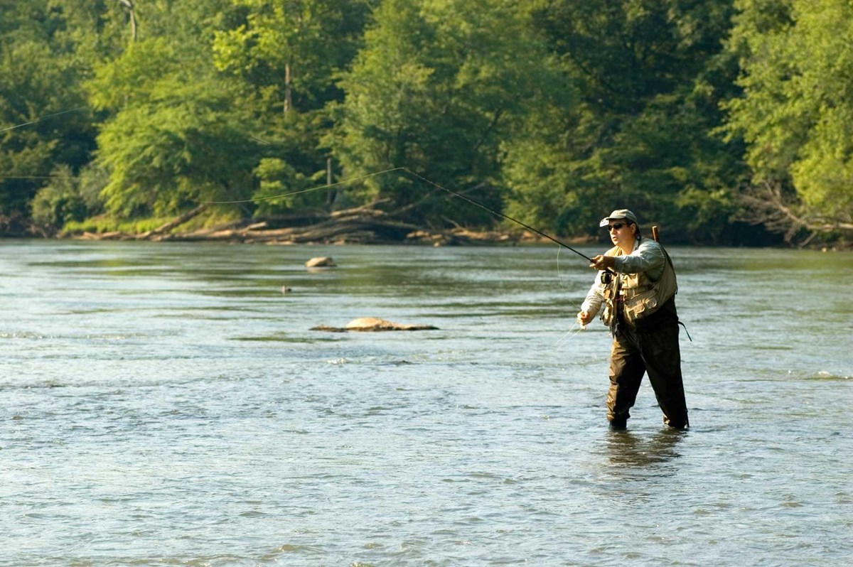 Angler in waders holding a fishing rod in the middle of a shallow river