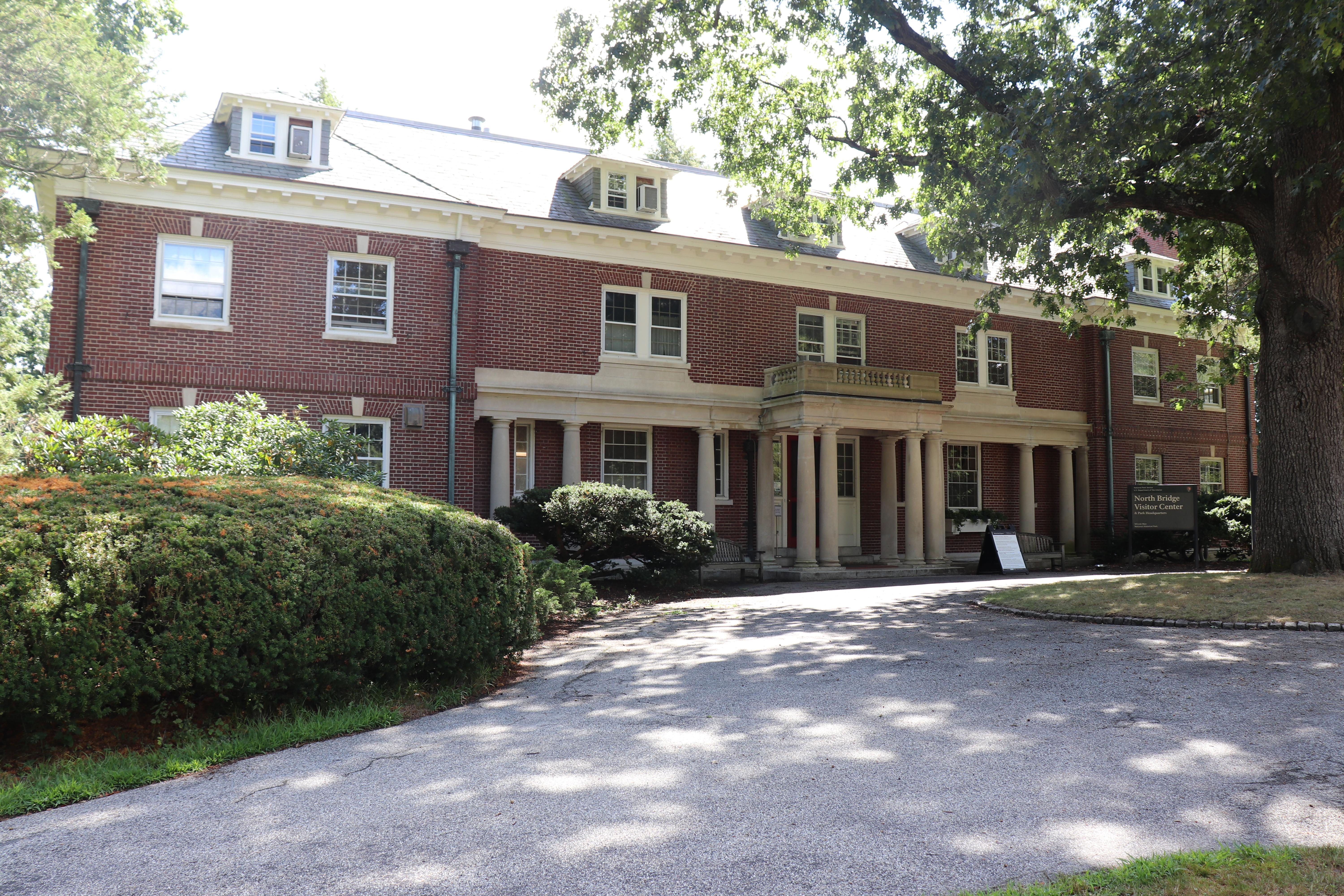 A three story brick mansion house with paved walkway leading to door. Green bushes flank both sides.