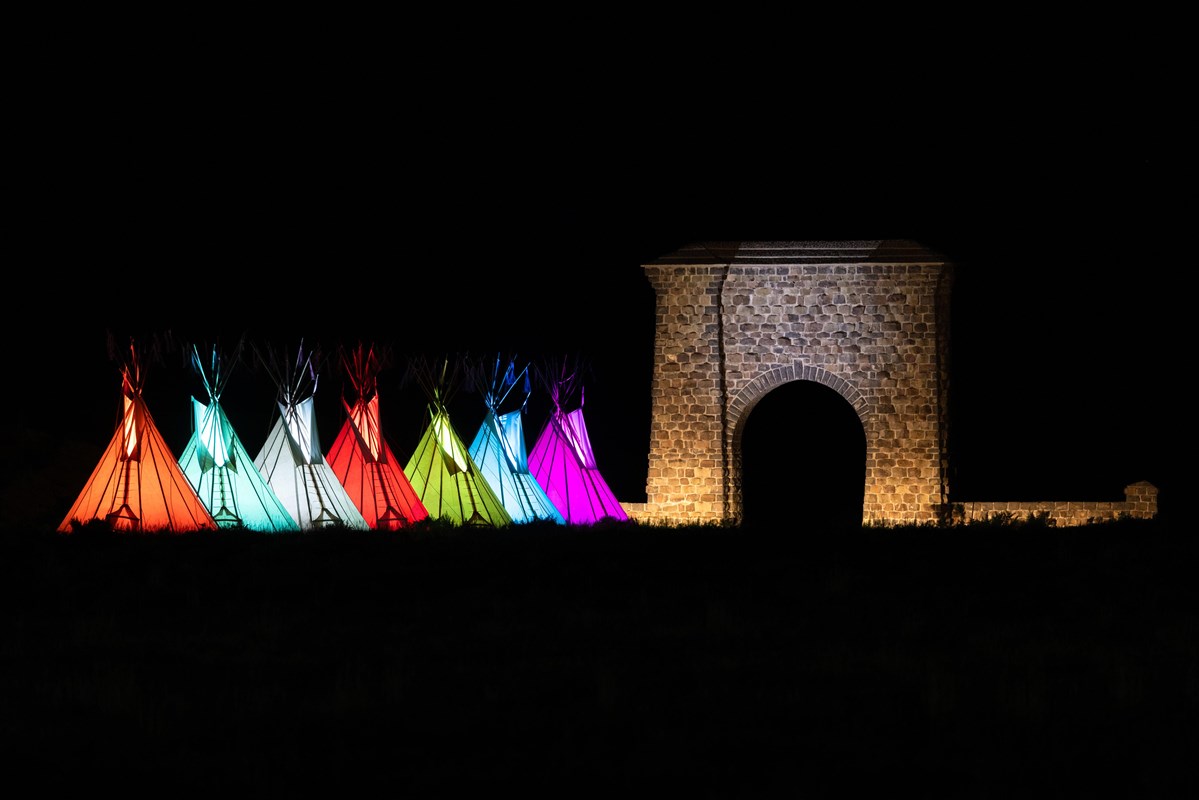 Multi-colored teepees are lit in front of a large, stone arch under the black night sky.