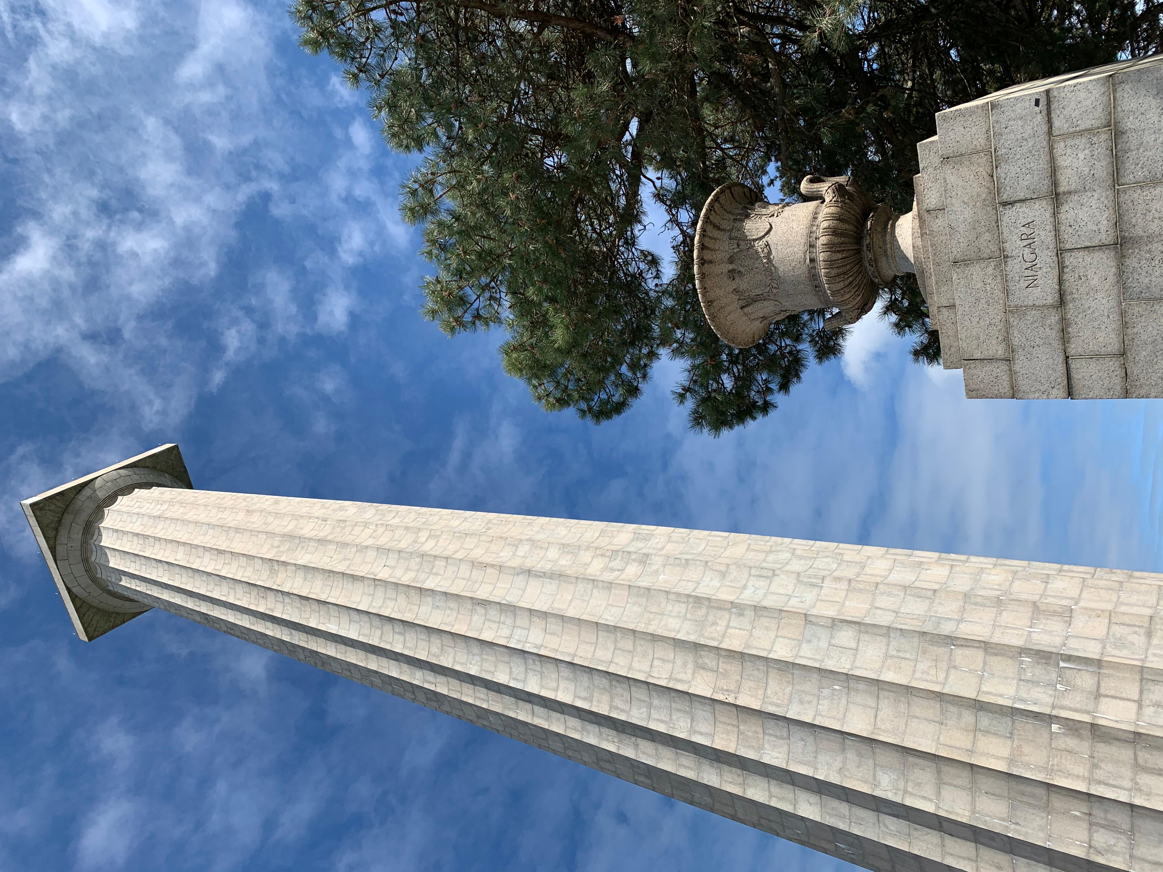 352 foot tall stone memorial column rises up to touch blue sky. In front a 5' urn on pedestal