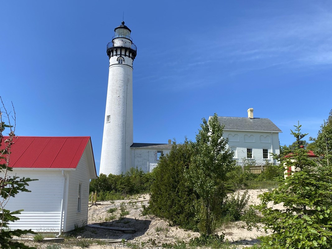 A white lighthouse next to a two story brick building with a red roofed building in the foreground.