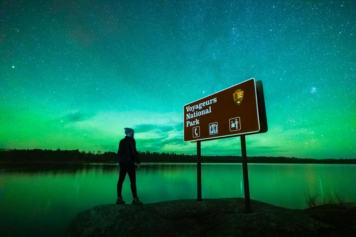 Photograph of person standing next to brown national park sign at night with northern lights. 