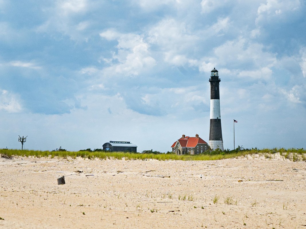 A wide shot of the white and black striped Fire Island Lighthouse from the beach. 