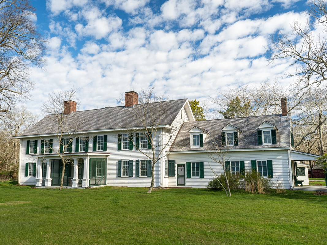 A white two-story colonial home with green shutters stands on a lawn with blue sky and white clouds.