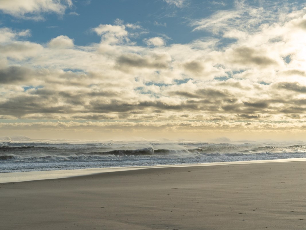 Waves crash on a sandy beach as the sun sets on a cloud-strewn sky.