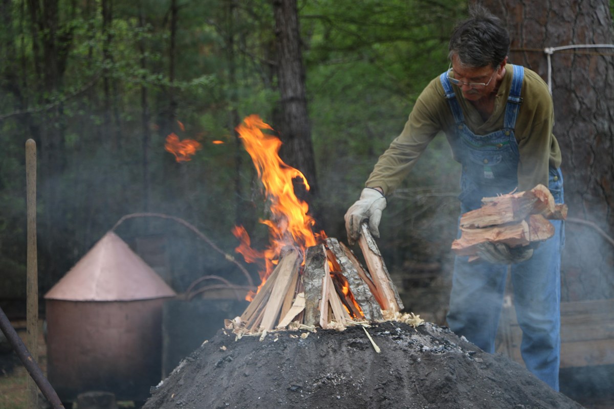 Man loading wood on to a dirt mound with a fire burning. 