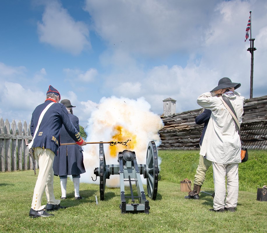 A cannon crew fires towards a wooden fort. An explosion of fire jets out in front of them. 