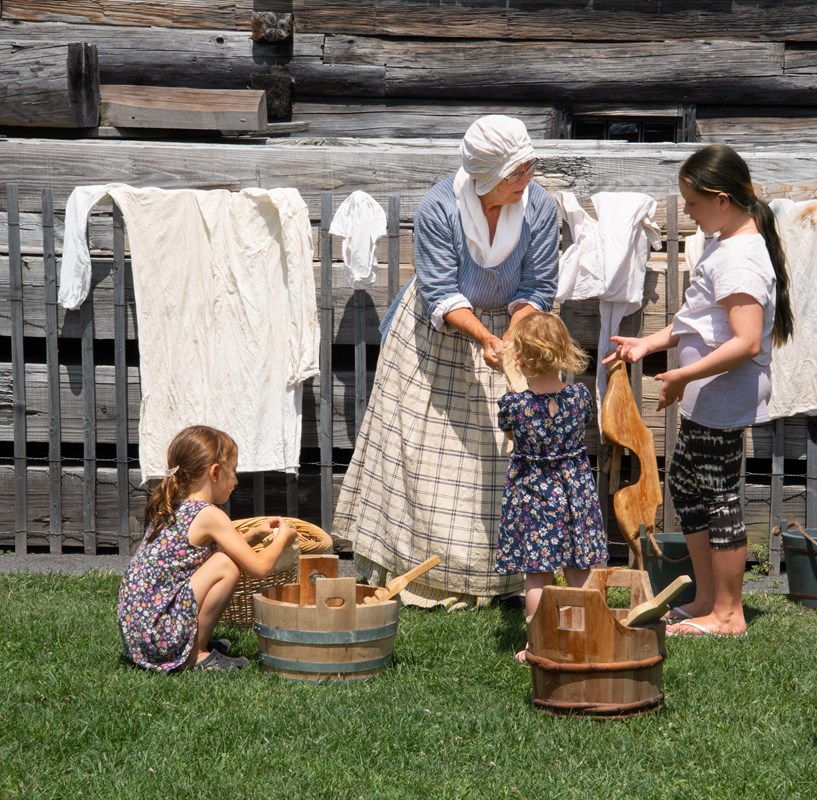 A woman in colonial clothes stands with a group of modern children, washing laundry in big buckets. 