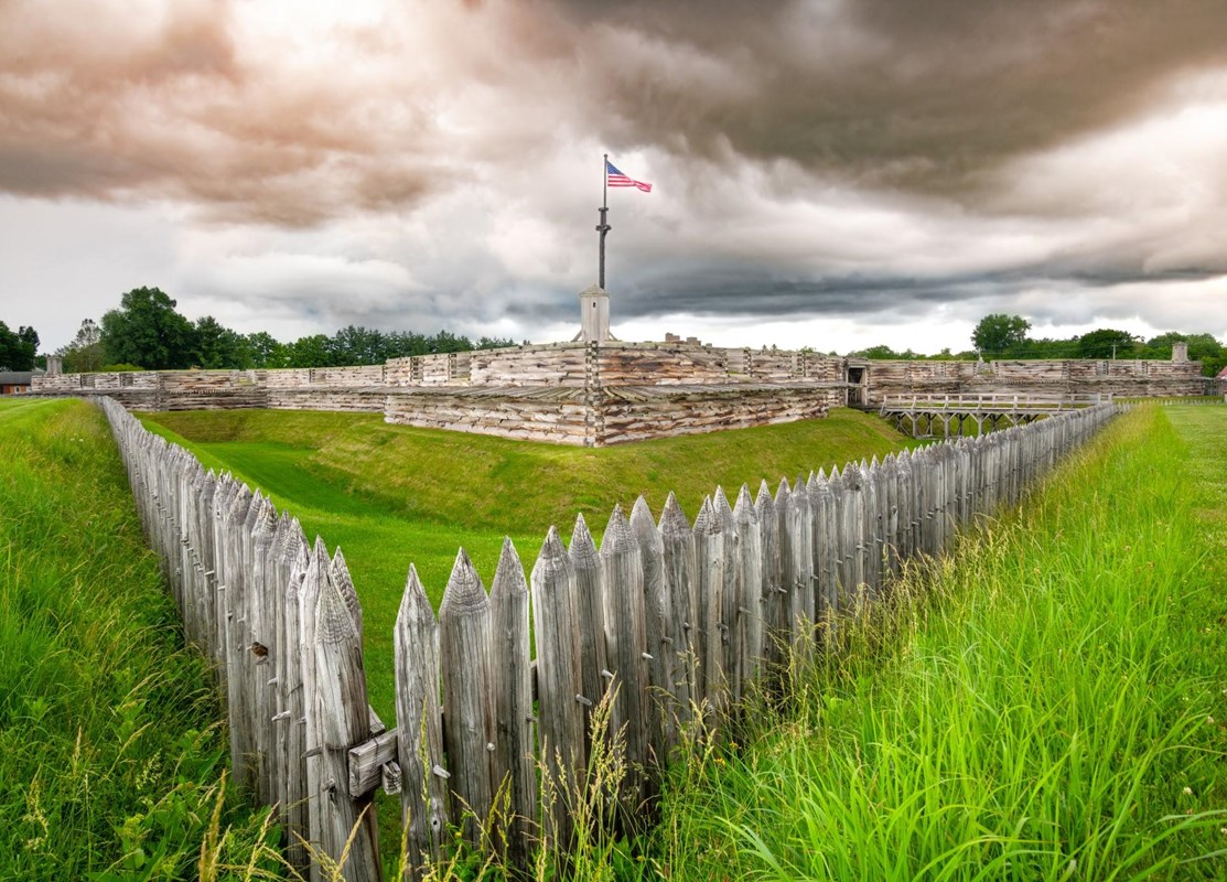 Puffy white clouds billow over log cabin-like walls. An American flag flies on a wooden pole