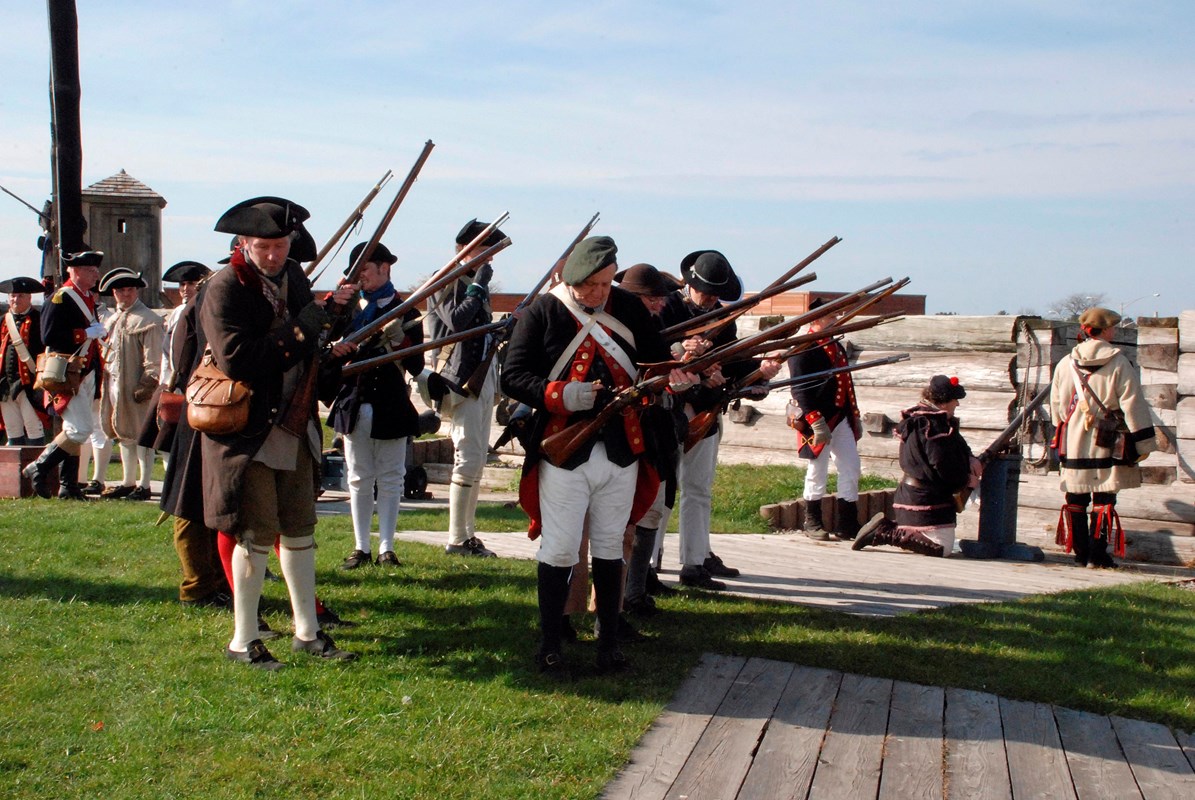 About two dozen soldiers stand in a neat rows holding their muskets.