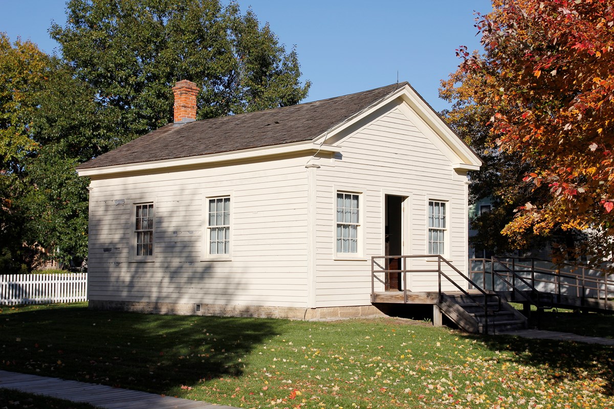 A one-story wooden building painted off-white has two windows and a central doorway.