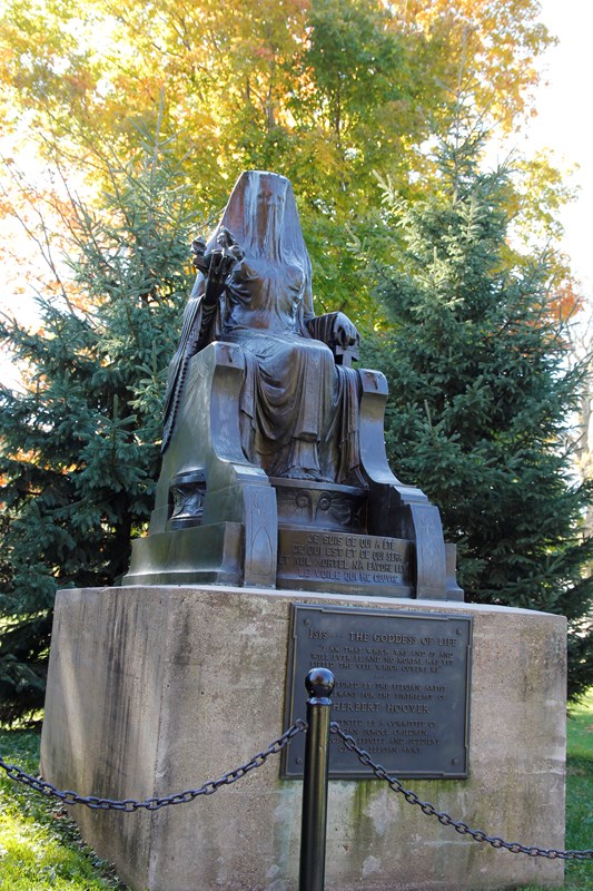 A cast bronze figure of a veiled goddess sits on a throne mounted on a concrete pedestal.