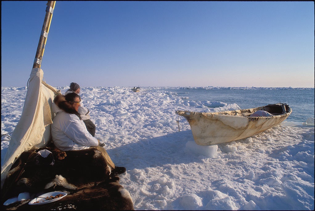 Two people sit by a tent and a skin canoe in snow next to the ocean.