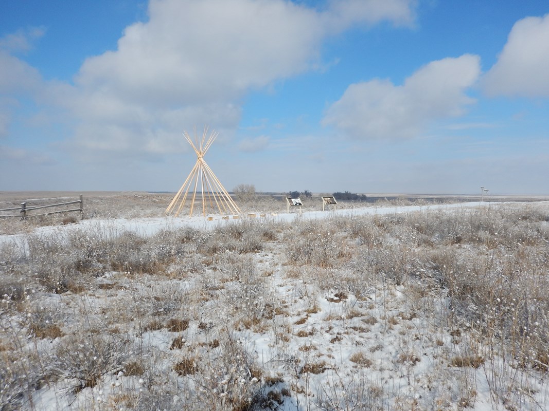 A lone tipi frame and two wayside signs stand in a snow-covered landscape 