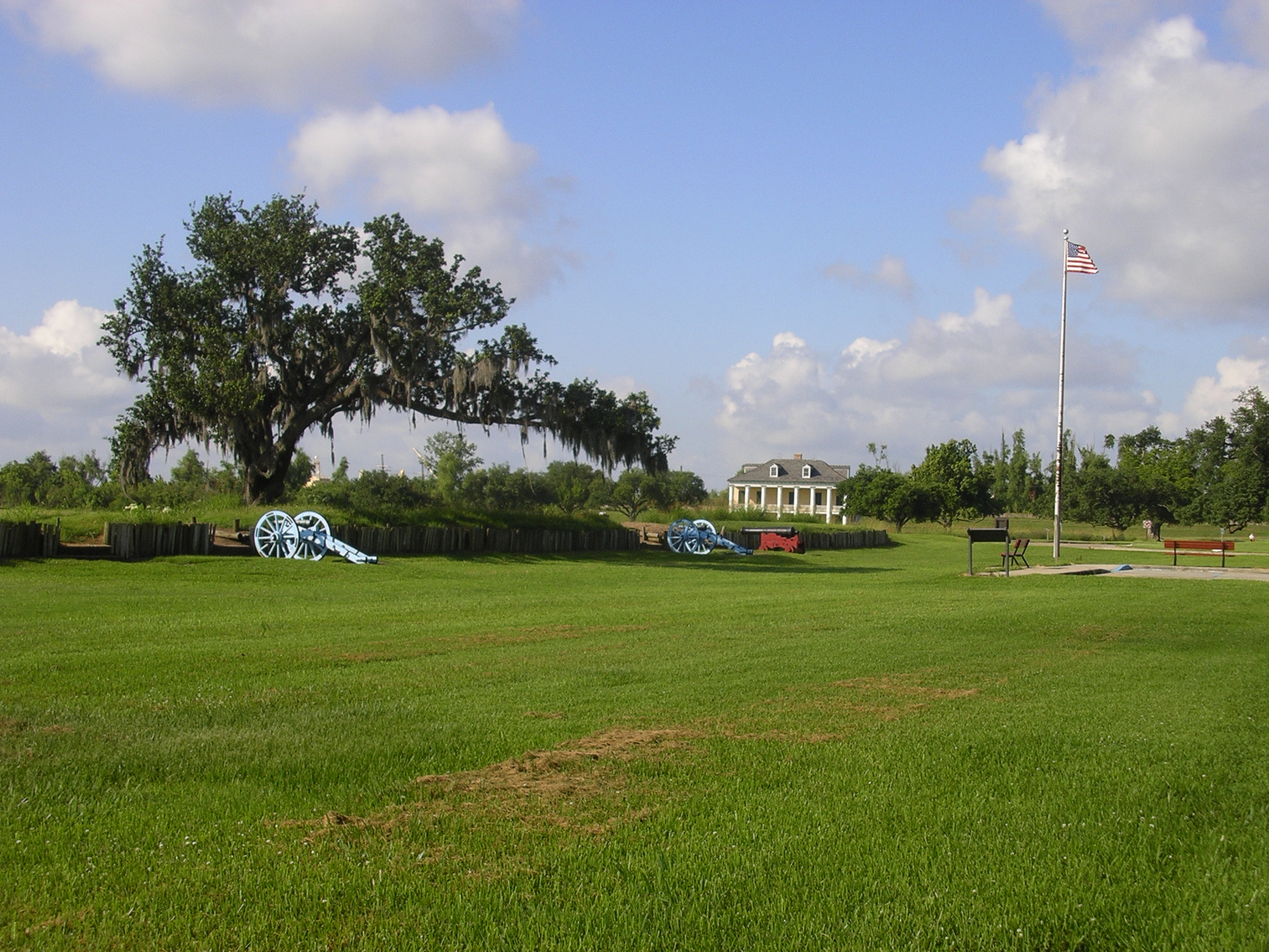 An oak tree arches over a cannon-guarded rampart with historic house and American flag in background