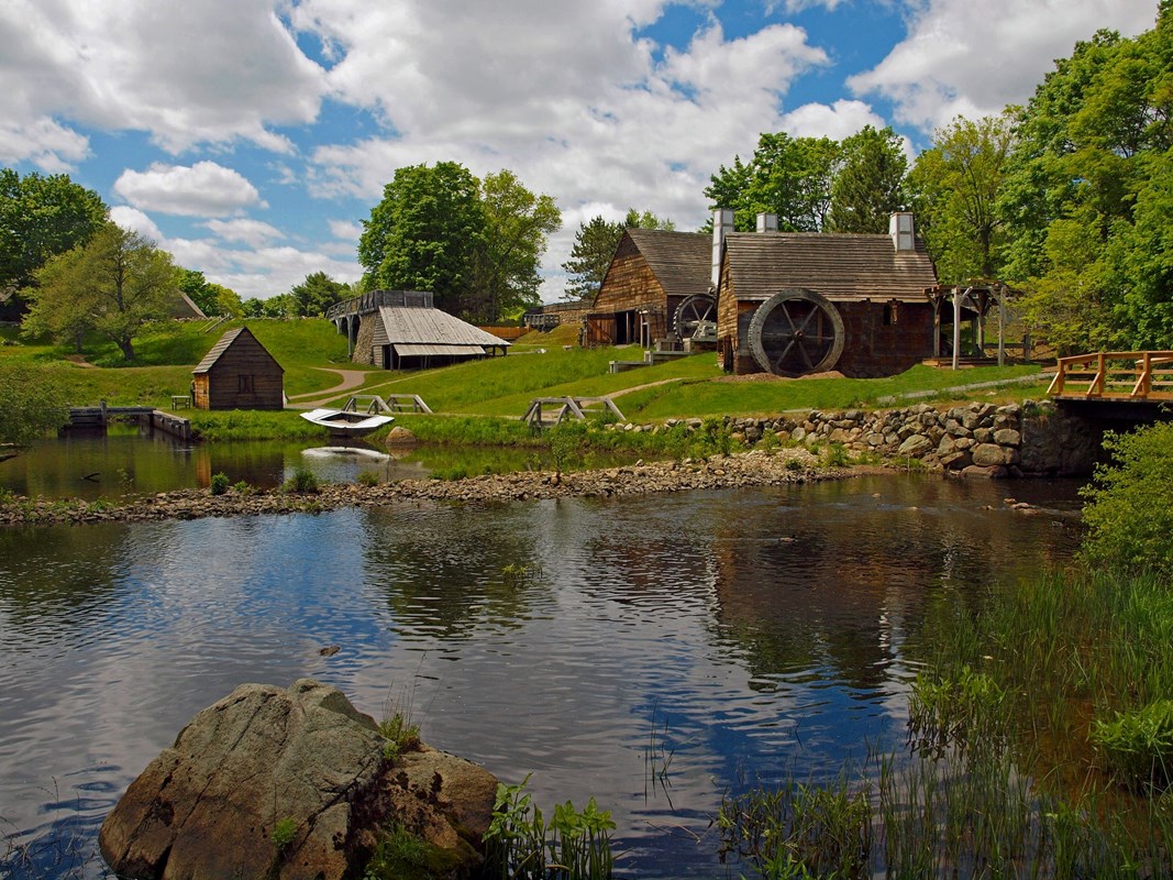 Several wooden structures amid green fields and trees under partly cloudy sky and beside river.