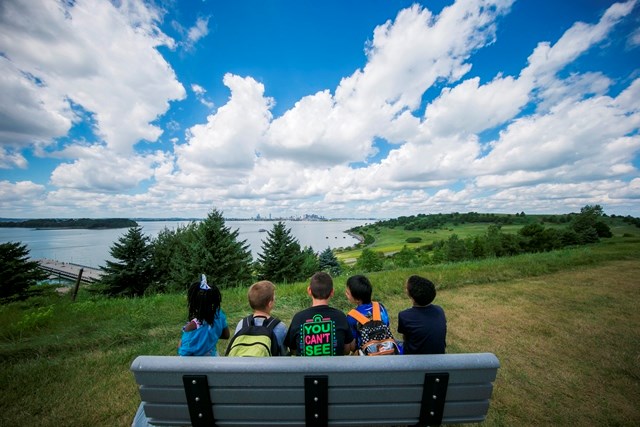 Group of children sit on a bench on a drumlin hill on Spectacle Island overlooking Boston Harbor
