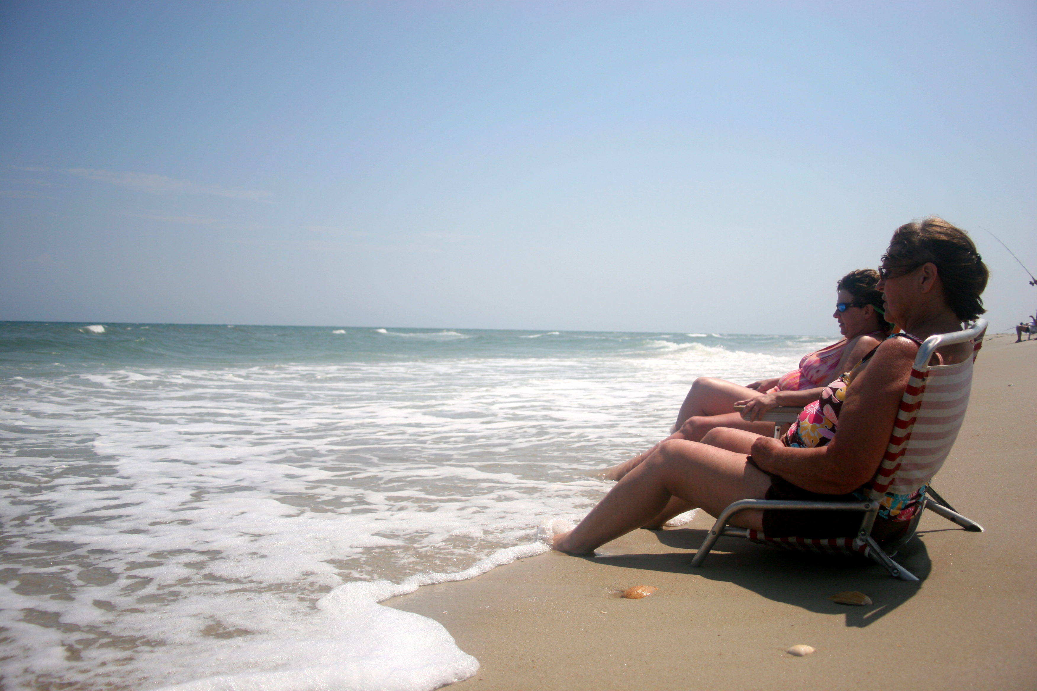 2 ladies sit in beach chairs on the sand with their feet in the water