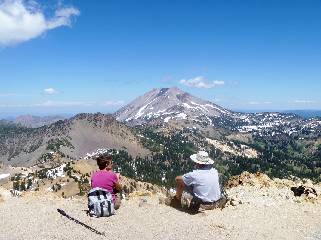 Two hikers sit on a mountain top with a view of multiple peaks, dotted with patches of snow 