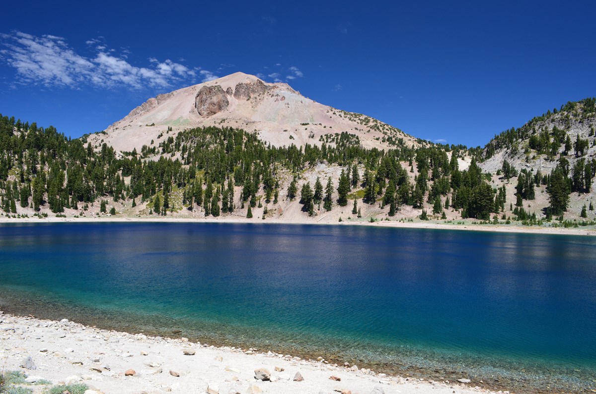 A volcanic peak rises above a tree-lined, deep blue lake. 