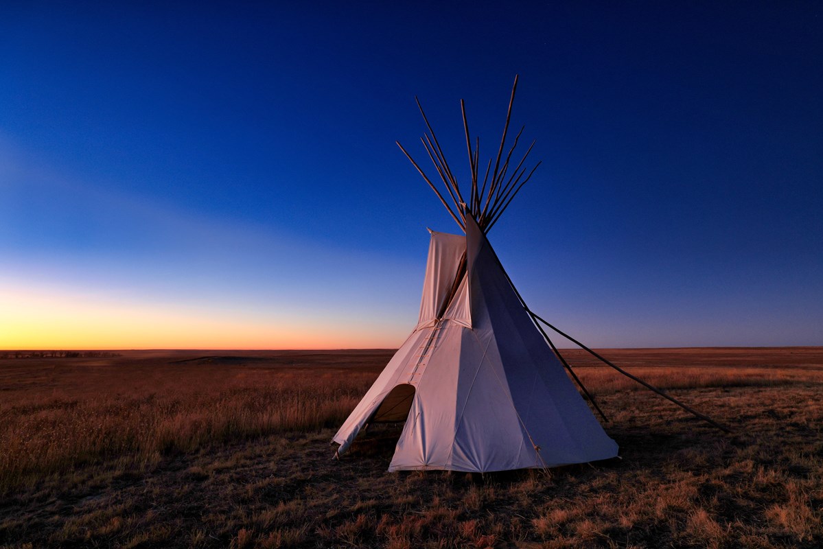 A lone tipi stands on a prairie landscape in predawn light. 