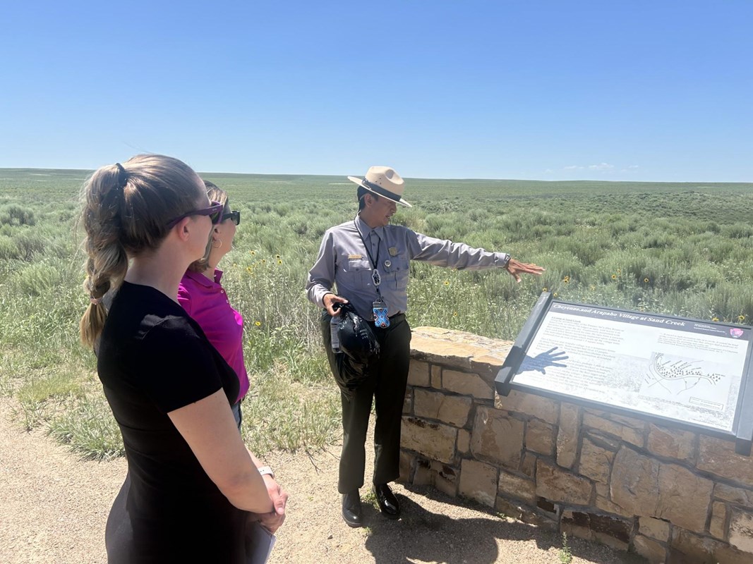 A park ranger points in the distance as two visitors stand before an outdoor exhibit panel. 