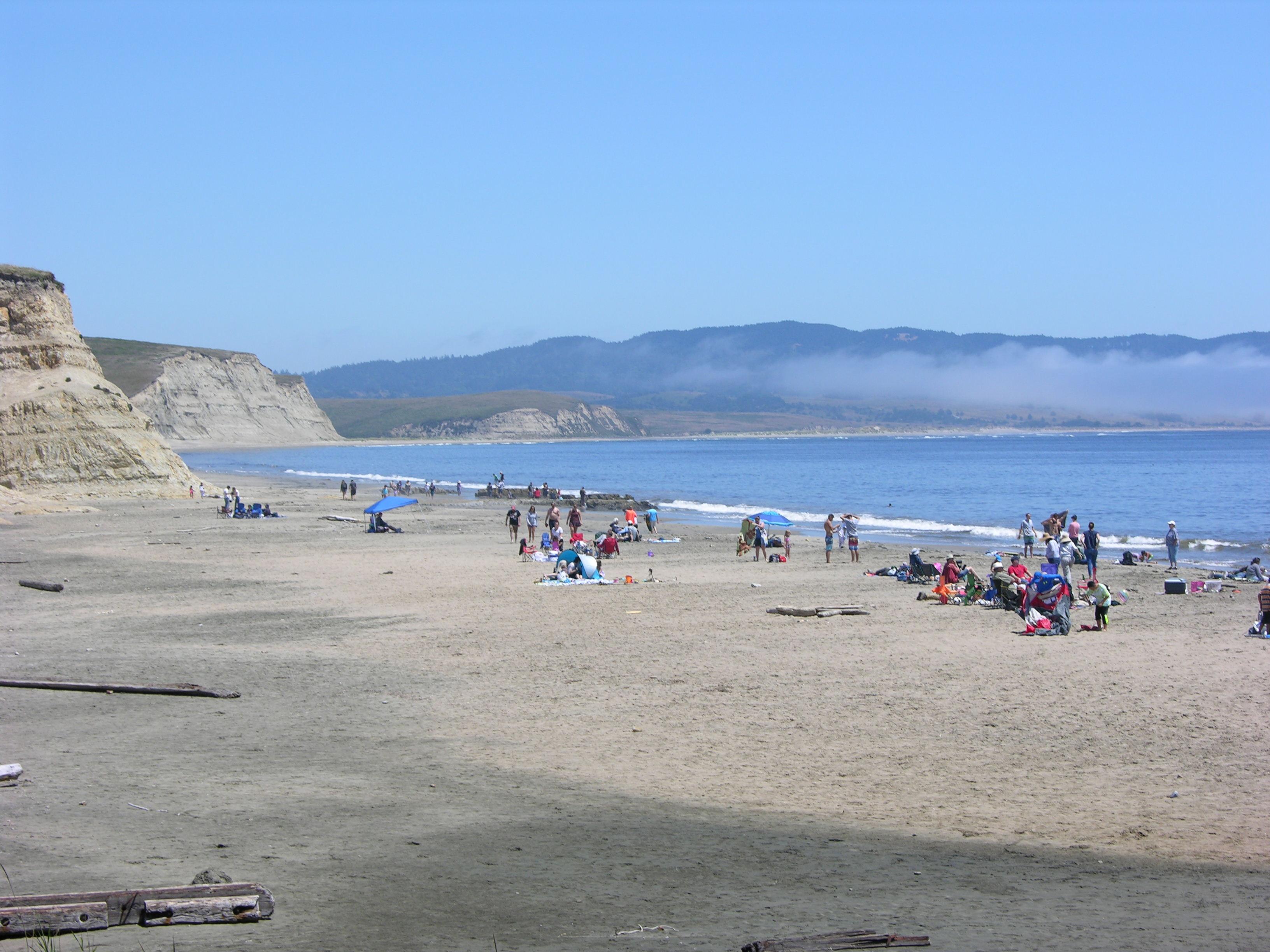 A few dozens of visitors walk along or sit on towels at a sandy beach on a sunny day.