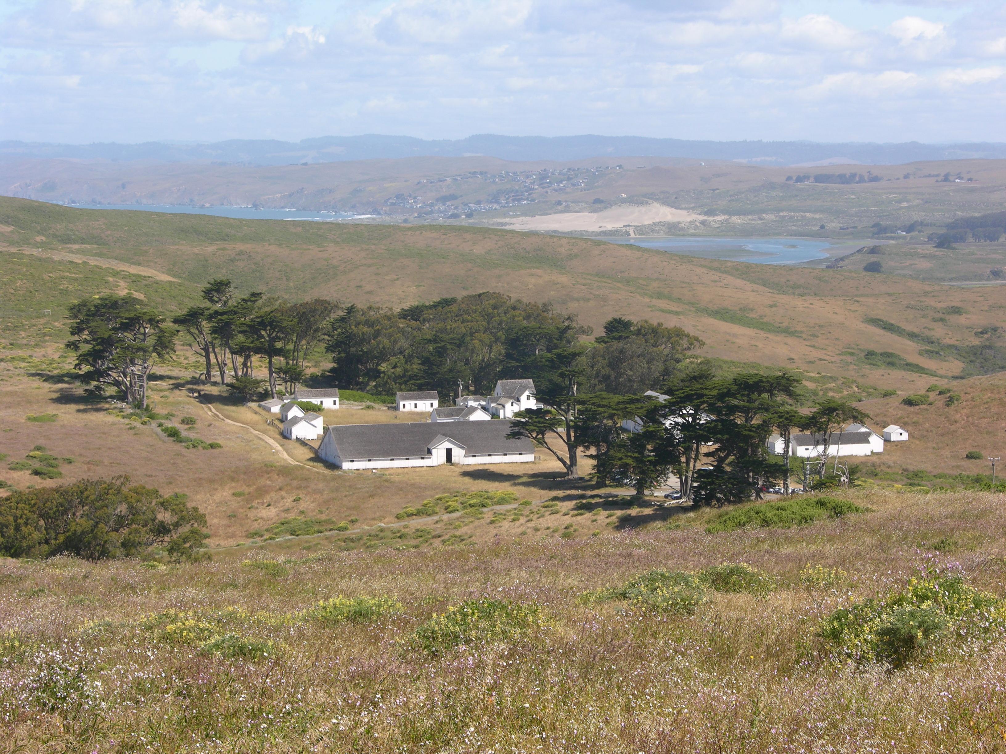 A historic dairy ranch composed of white-painted buildings surrounded by dry grass and a few trees.