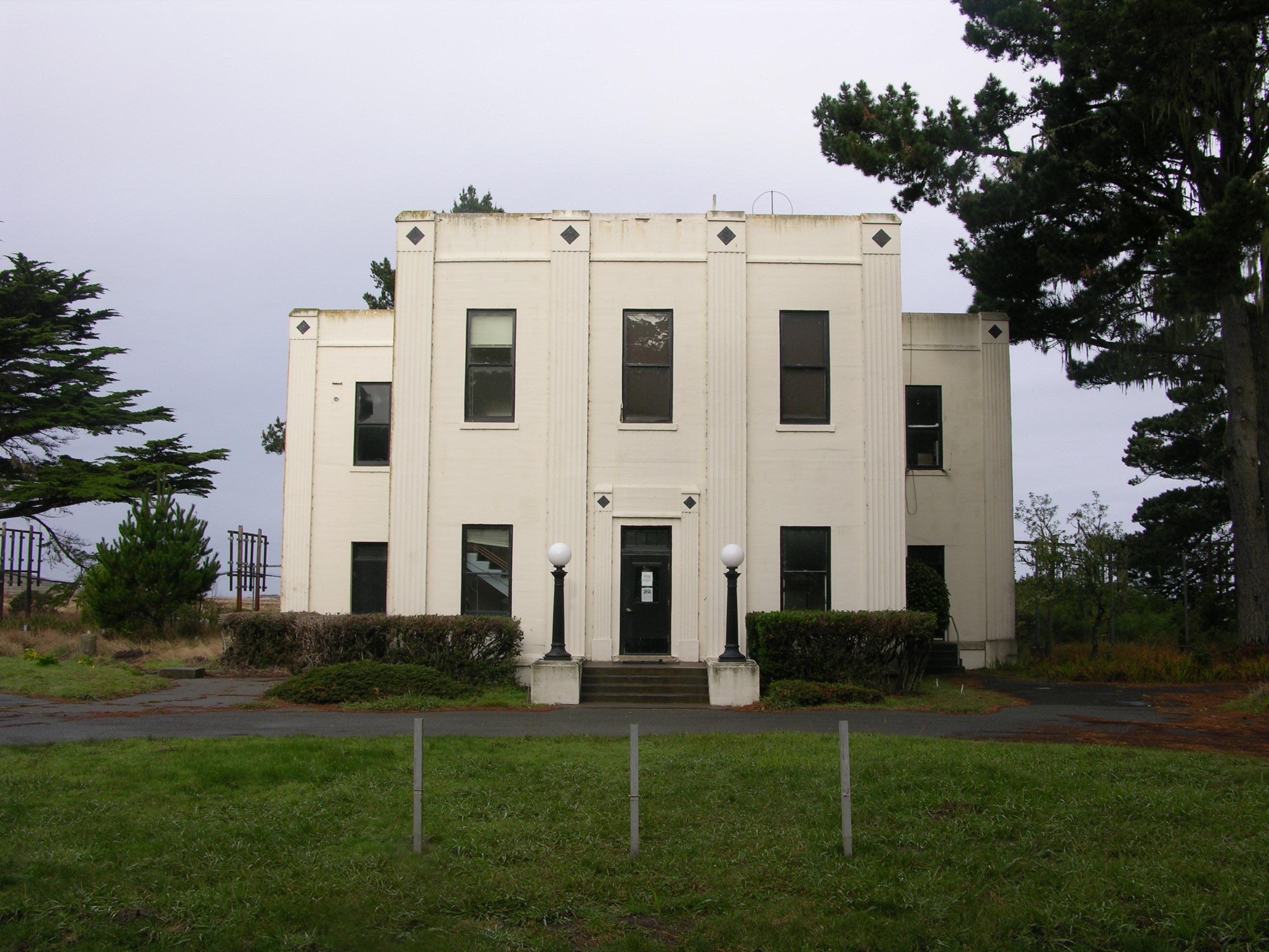 A white, two-story art deco building beyond a green, grassy round-about.