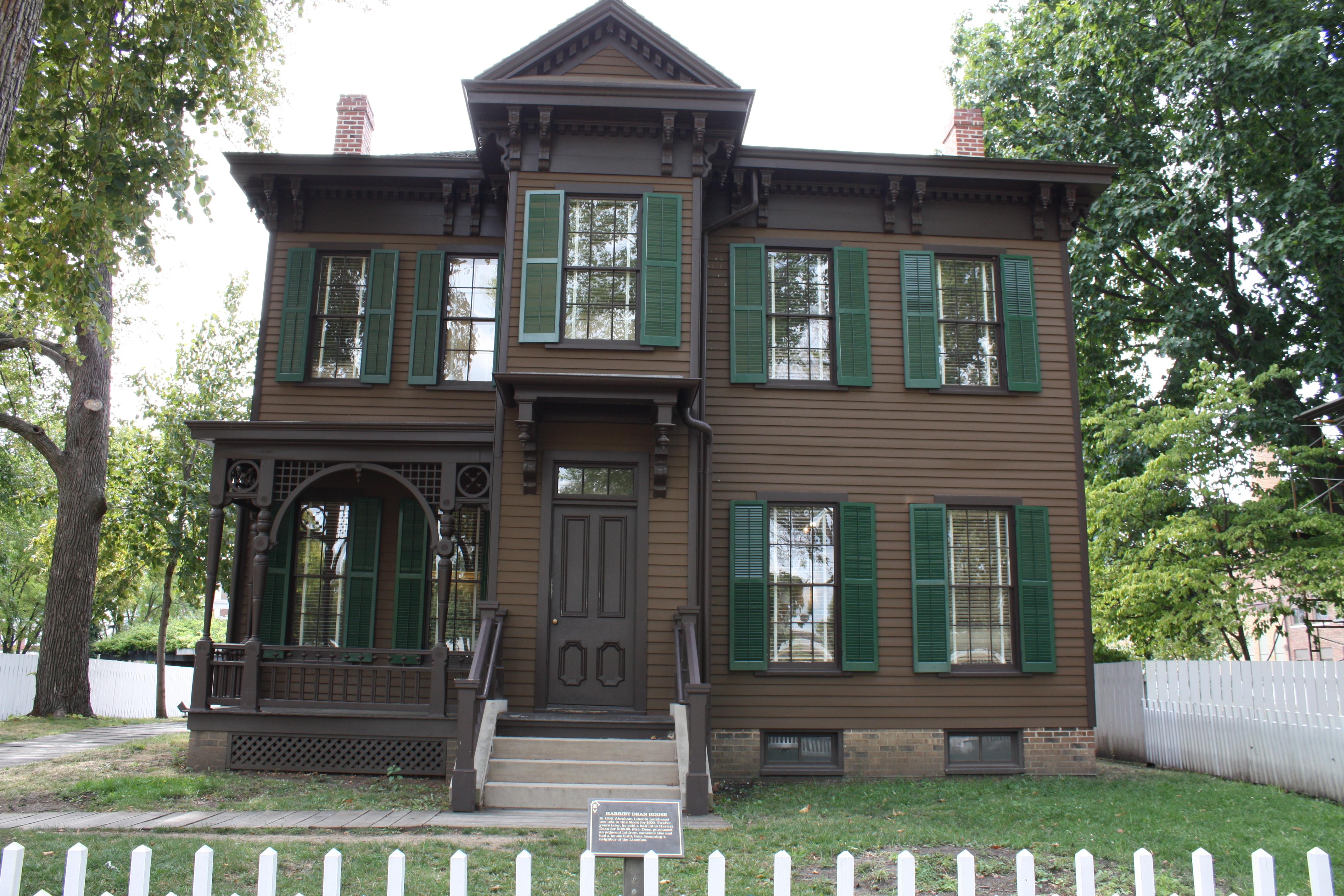 2 story brown house with dark green shutters and white fence.