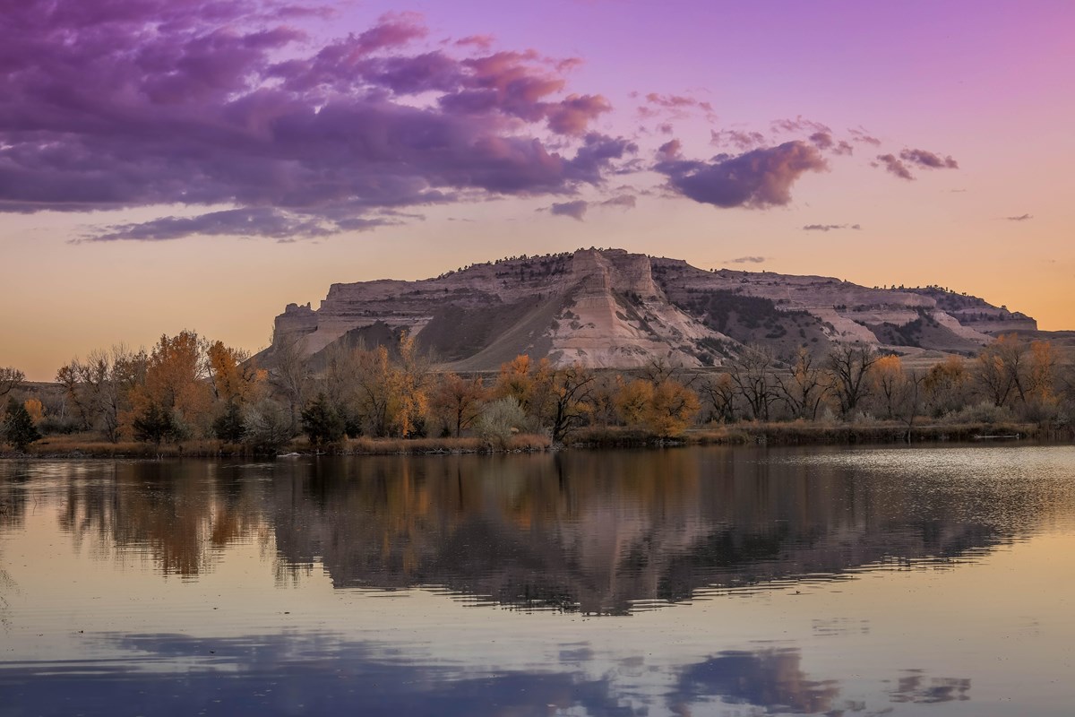 A pink sky and dramatic bluff are seen reflected in water.