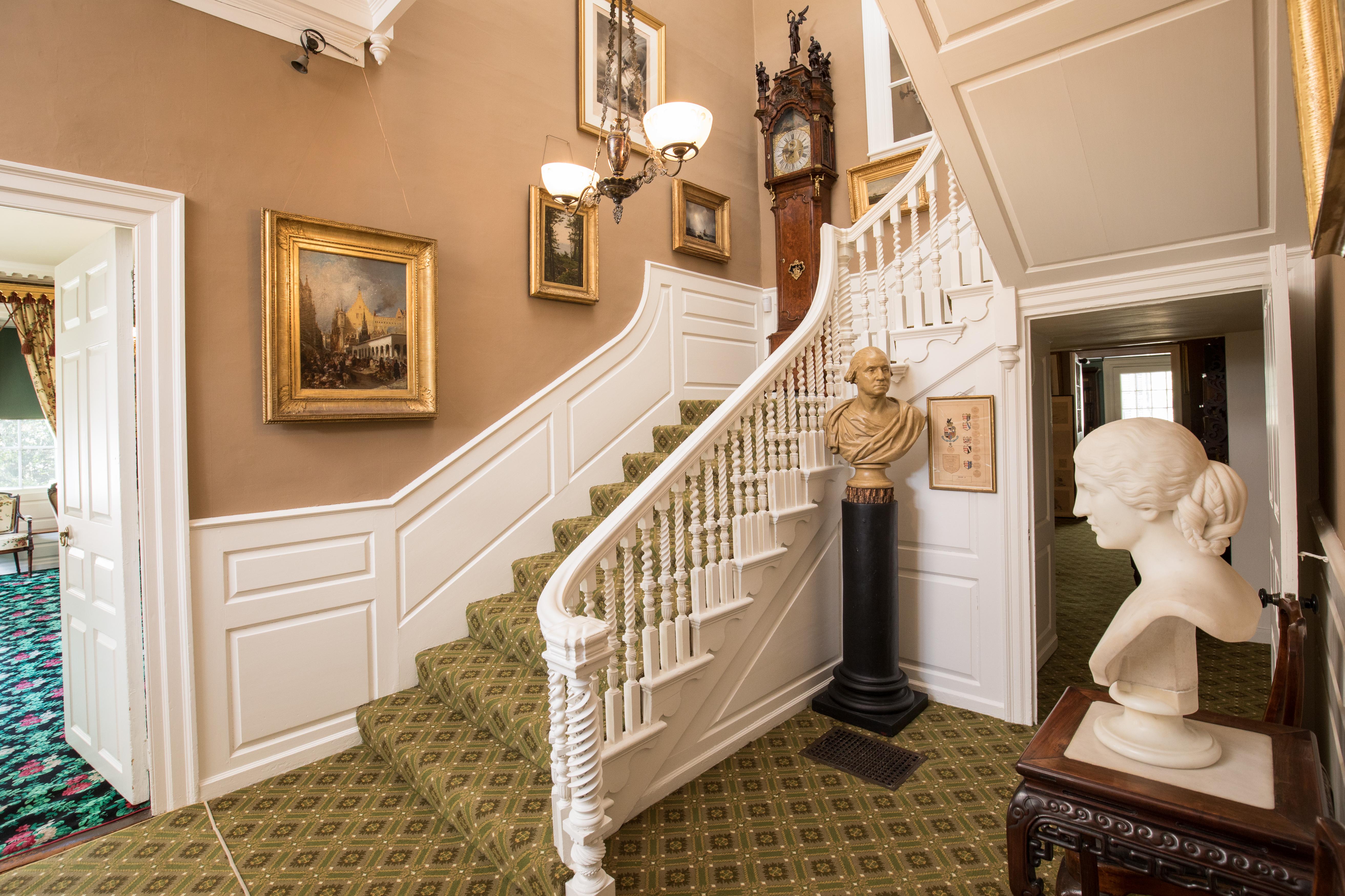 A staircase with white balusters and green carpet. A bust of Washington sits at the turn of stairs.