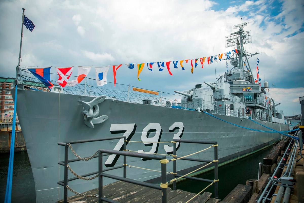 A gray naval destroyer sits next to a dock. It has "793" on the side and colorful signal flags.