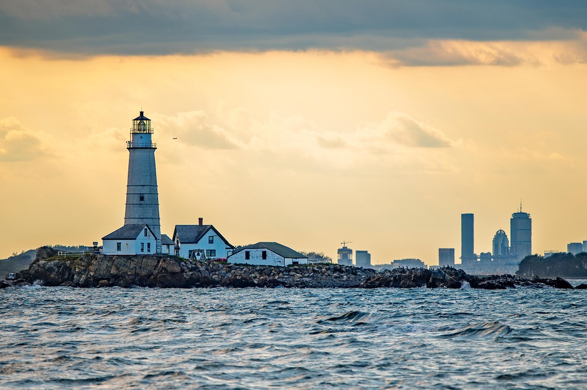 a white lighthouse and other buildings on Little Brewster Island at sunset. City in the distance.