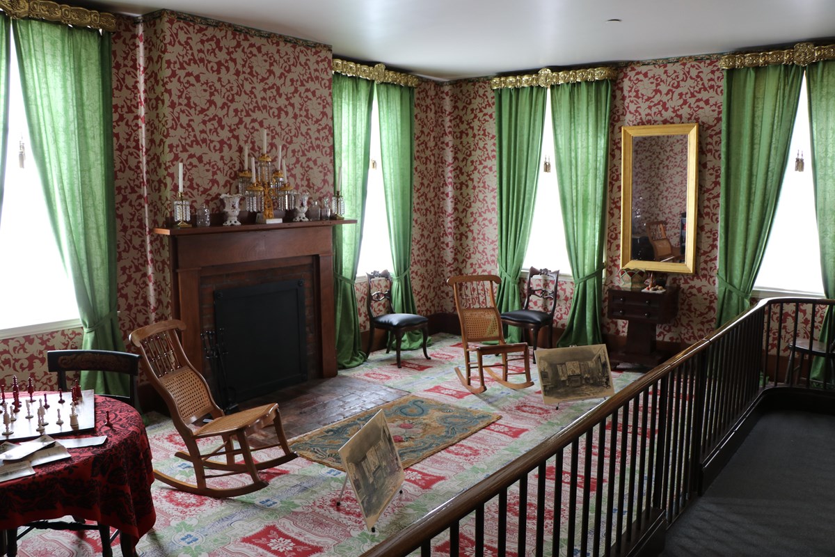 A room with patterned carpet and wallpaper, a fireplace, several wooden chairs, and decorations.