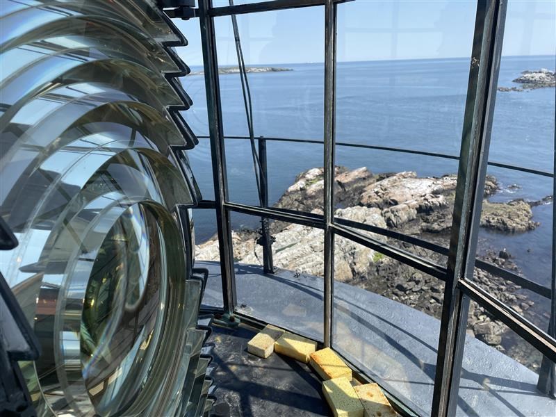 A lighthouse lens to the left, and to the right is a window looking out over a rocky shoreline.