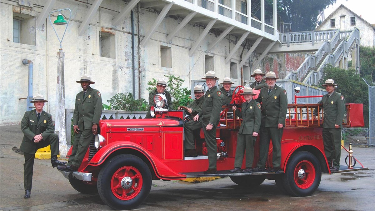 Ten people in green uniforms and flat brimmed hats stand on and around a red antique fire truck