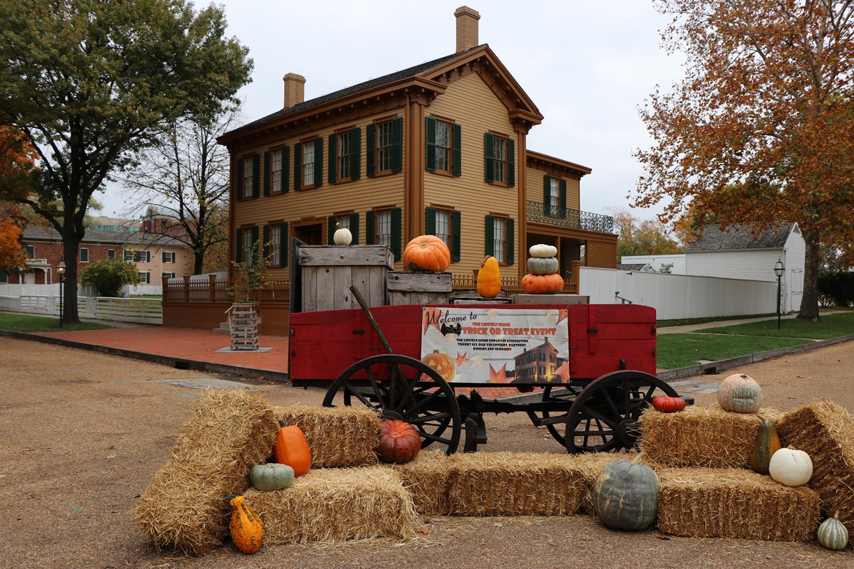A red cart surrounded by pumpkins and hay bales with the Lincoln Home in the background.